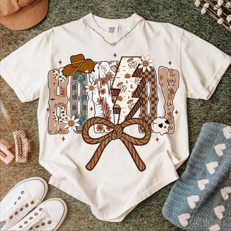 May include: Cream-coloured t-shirt with the word "HONEY" in a decorative font, featuring a cowboy hat, flowers, and a lightning bolt design. A brown rope bow is at the bottom. The shirt is surrounded by accessories.