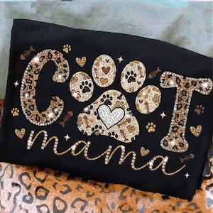 May include: Black t-shirt with the text "Cat Mama" in a sparkling design. The letters "C" and "T" have a leopard print pattern, and the paw prints and hearts have a similar design. The word "mama" is in a cursive font.
