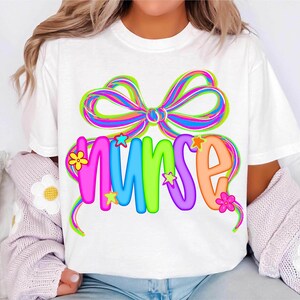 May include: White t-shirt with the word "NURSE" in bright, bubble letters, decorated with a rainbow bow and star and flower details. The design is colourful and appealing, suitable for healthcare workers.