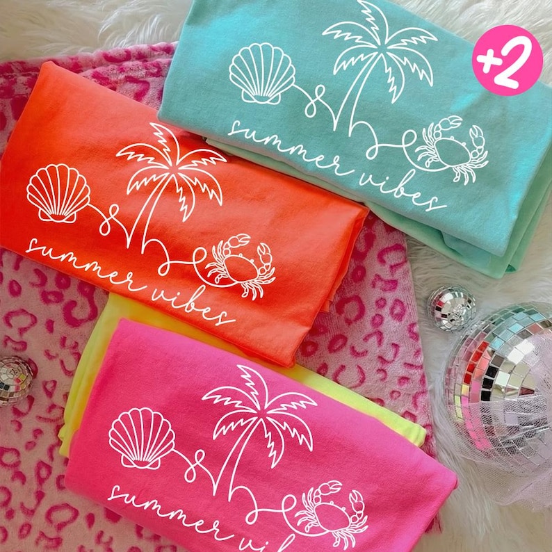May include: Four folded t-shirts in bright colors: orange, pink, teal, and yellow. Each shirt features a white graphic of a seashell, palm tree, and crab, with the words "summer vibes" in a script font. A pink circle with "+2" is in the top right corner.