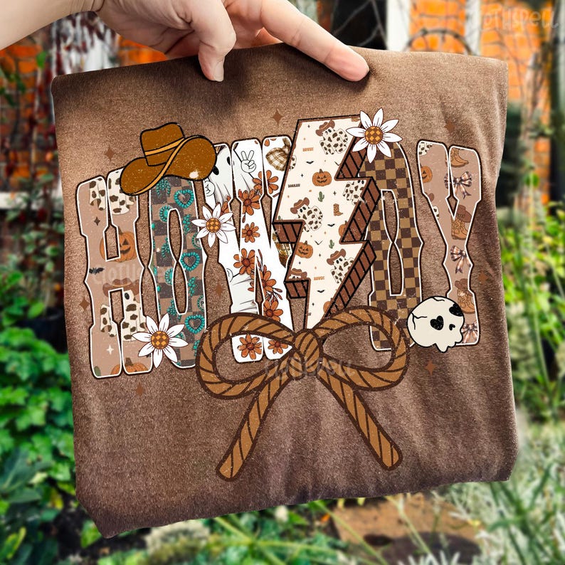 May include: Brown t-shirt featuring the word "HONEY" in a decorative font. The design includes a cowboy hat, lightning bolt, flowers, and a skull. The shirt has a large brown rope bow at the bottom.