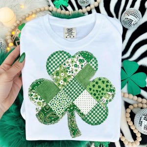 Puede incluir: Camiseta blanca de cuello redondo con un dise&ntilde;o de tr&eacute;bol de retazos. El tr&eacute;bol est&aacute; compuesto por varias telas verdes estampadas, incluyendo patrones florales, de lunares y a cuadros. La camiseta tiene una etiqueta que dice "Comfort Colors".