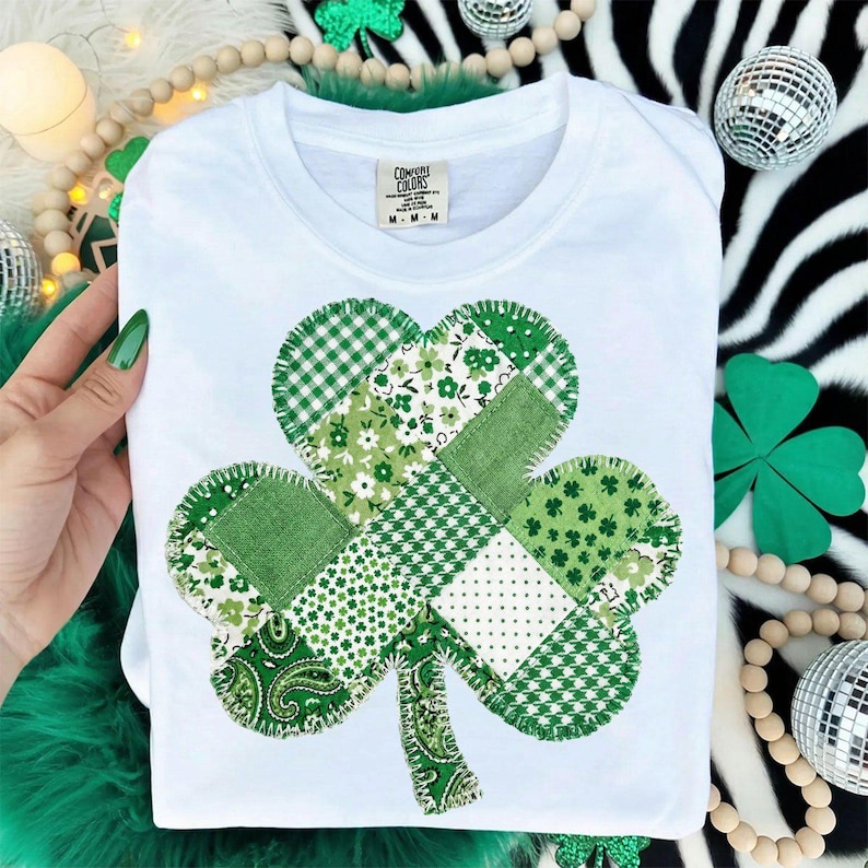 Puede incluir: Camiseta blanca de cuello redondo con un dise&ntilde;o de tr&eacute;bol de retazos. El tr&eacute;bol est&aacute; compuesto por varias telas verdes estampadas, incluyendo patrones florales, de lunares y a cuadros. La camiseta tiene una etiqueta que dice "Comfort Colors".