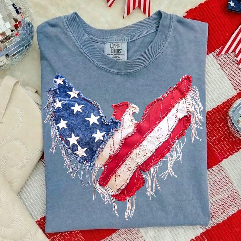 May include: A blue Comfort Colors t-shirt featuring an American flag eagle design. The eagle's wings are crafted from denim and red fabric, with white stars and stripes. The design has a distressed, fringed edge.