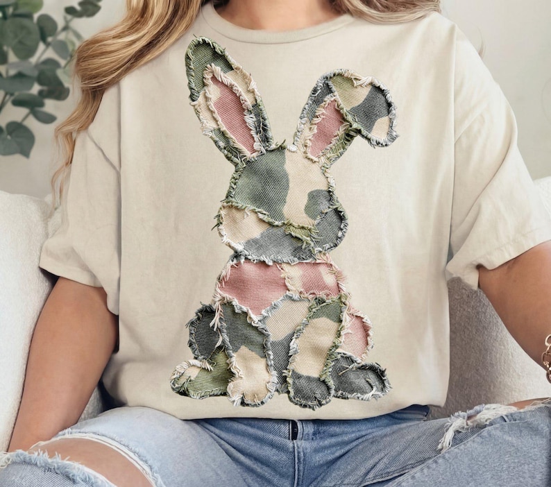 May include: Cream-colored t-shirt featuring a patchwork bunny design. The bunny is made of various fabric pieces in shades of green, pink, and beige, with a distressed, frayed edge. The shirt has short sleeves and a relaxed fit.