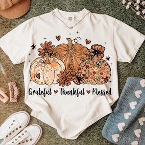 May include: Cream-colored t-shirt featuring a fall-themed design. The graphic includes three pumpkins with floral and leopard print patterns, surrounded by flowers and hearts. The text "Grateful Thankful Blessed" is printed below the pumpkins.