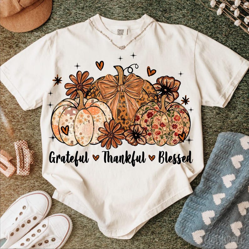 May include: Cream-colored t-shirt featuring a fall-themed design. The graphic includes three pumpkins with floral and leopard print patterns, surrounded by flowers and hearts. The text "Grateful Thankful Blessed" is printed below the pumpkins.
