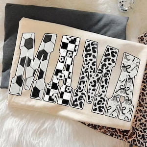 May include: A cream-colored t-shirt with the word "MAMA" in large, bold letters. The letters are decorated with soccer ball, checkered, leopard print, and bow patterns. The shirt is laying on a white fluffy surface.