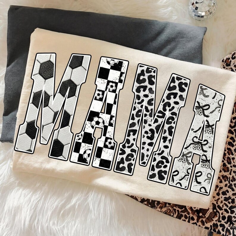 May include: A cream-colored t-shirt with the word "MAMA" in large, bold letters. The letters are decorated with soccer ball, checkered, leopard print, and bow patterns. The shirt is laying on a white fluffy surface.
