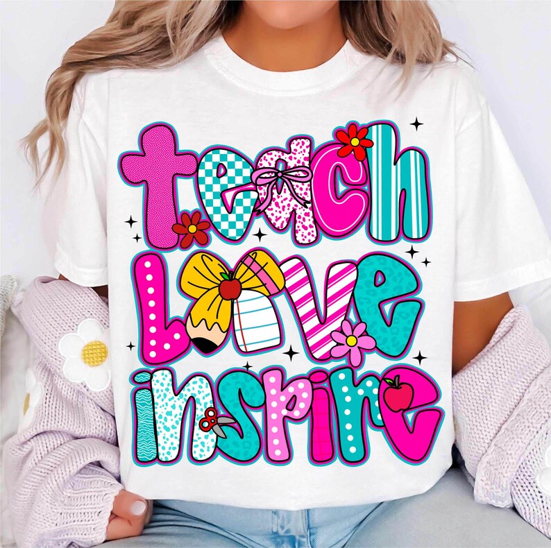 May include: White t-shirt with the words "Teach Love Inspire" in colorful, playful lettering. The design incorporates flowers, a pencil, and a bow, with a mix of patterns and colors, including pink, teal, and yellow.