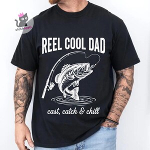 May include: Black t-shirt featuring the text "REEL COOL DAD" above a white illustration of a fish leaping from water, with a fishing rod. The words "cast, catch & chill" are below the fish graphic.