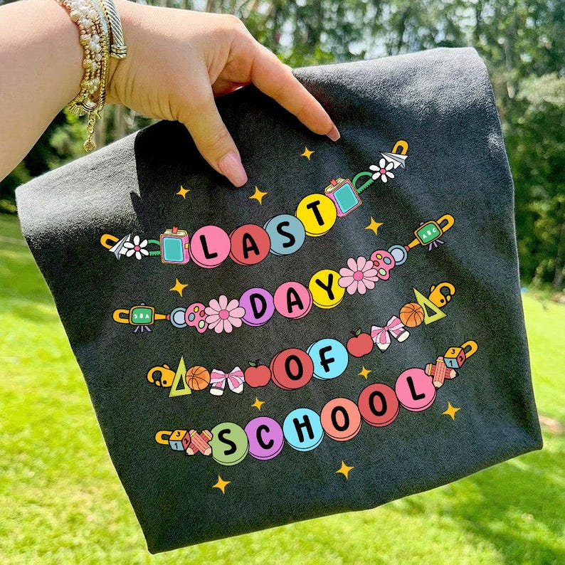 May include: A dark gray t-shirt with the words "LAST DAY OF SCHOOL" in colorful bubble letters. The text is arranged in three rows, each adorned with decorative elements like flowers, apples, and stars. The shirt is held up by a person.