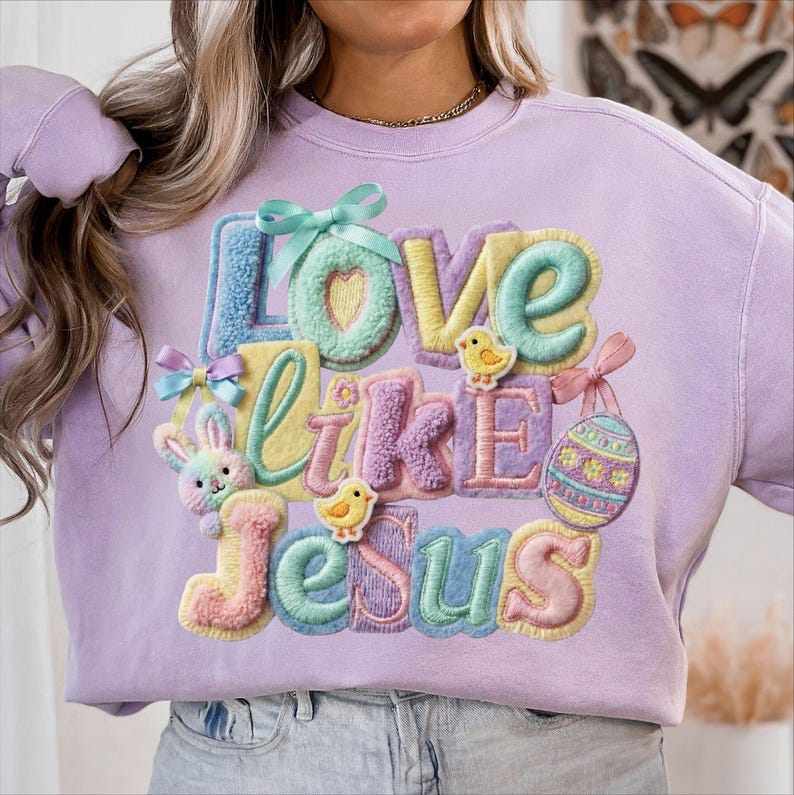 Pu&ograve; includere: Una felpa lilla con la scritta "Love Like Jesus" in lettere colorate e strutturate. Il design include elementi pasquali come un coniglietto, pulcini e un uovo di Pasqua, il tutto in colori pastello.