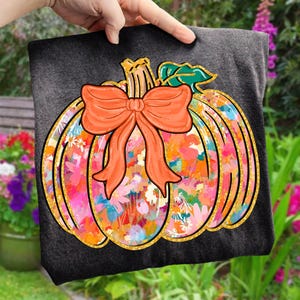 May include: Dark gray t-shirt featuring a colorful pumpkin design. The pumpkin has a floral pattern in shades of pink, orange, and blue, outlined with gold glitter. An orange bow adorns the top. The design is perfect for fall and autumn.