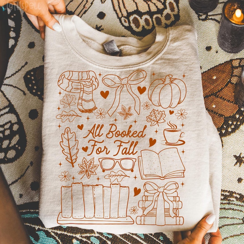 May include: Cream-colored sweatshirt with a fall-themed graphic. The design includes a scarf, bow, pumpkin, leaves, coffee cup, glasses, books, and the text "All Booked For Fall" in brown. The sweatshirt is folded.