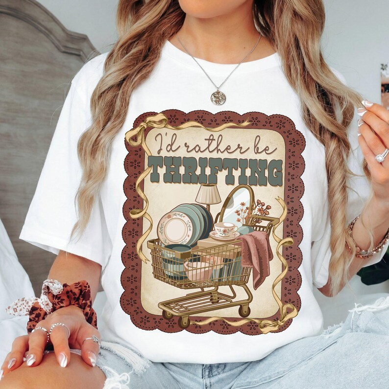 May include: White t-shirt with a graphic featuring a shopping cart filled with vintage items, including plates, a lamp, and a mirror. The text "I'd rather be thrifting" is displayed above the cart in a vintage font.