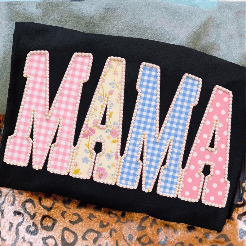 May include: Black shirt with the word "MAMA" in large, colorful letters. The letters are decorated with pink gingham, floral, blue gingham, and pink polka dot patterns, outlined with small white pearls.