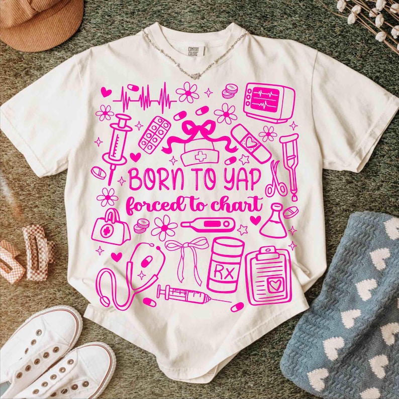 May include: Off-white t-shirt featuring a pink medical-themed design. The graphic includes medical symbols and the text "BORN TO YAP forced to chart." Other items, such as shoes and a hat, are visible around the shirt.