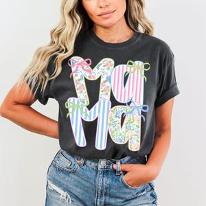 May include: Dark gray t-shirt with the word "MAMA" in large, colorful letters. The letters are decorated with floral patterns, pink and blue stripes, and bows. The shirt is a casual style, suitable for everyday wear.