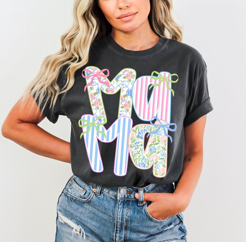 May include: Dark gray t-shirt with the word "MAMA" in large, colorful letters. The letters are decorated with floral patterns, pink and blue stripes, and bows. The shirt is a casual style, suitable for everyday wear.