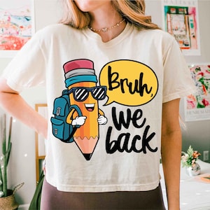 May include: Cream-colored t-shirt featuring a cartoon pencil character wearing sunglasses and a backpack. The pencil is yellow, orange, and pink. Text in a yellow speech bubble says "Bruh" and the text "we back" is below.