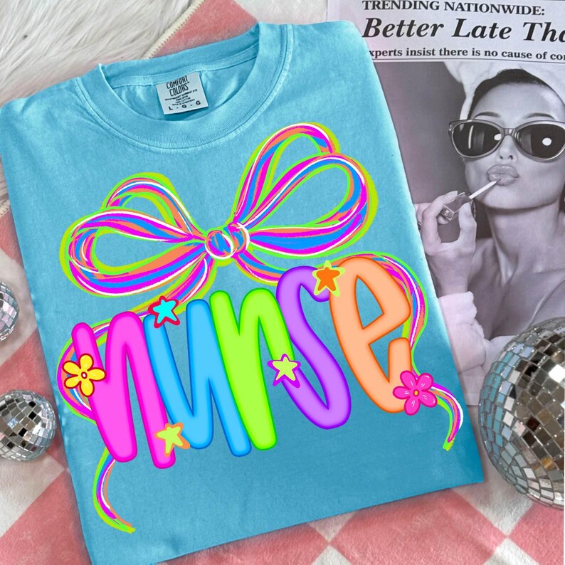 May include: A turquoise t-shirt featuring a large, multi-coloured bow and the word "NURSE" in bright, bubble letters. The design incorporates stars and flowers, giving a cheerful appearance. The shirt is a Comfort Colors brand.