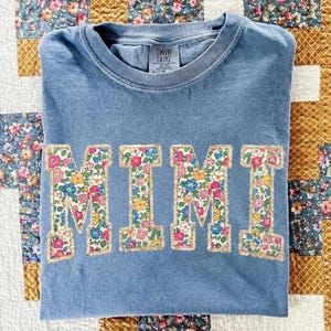 K&ouml;nnte beinhalten: Blaues Comfort Colors T-Shirt mit dem Wort "MIMI" in gro&szlig;en Buchstaben mit Blumenmuster und goldenem Rand. Das T-Shirt liegt gefaltet auf einem floralen Quilt. Ein l&auml;ssiges Kleidungsst&uuml;ck.