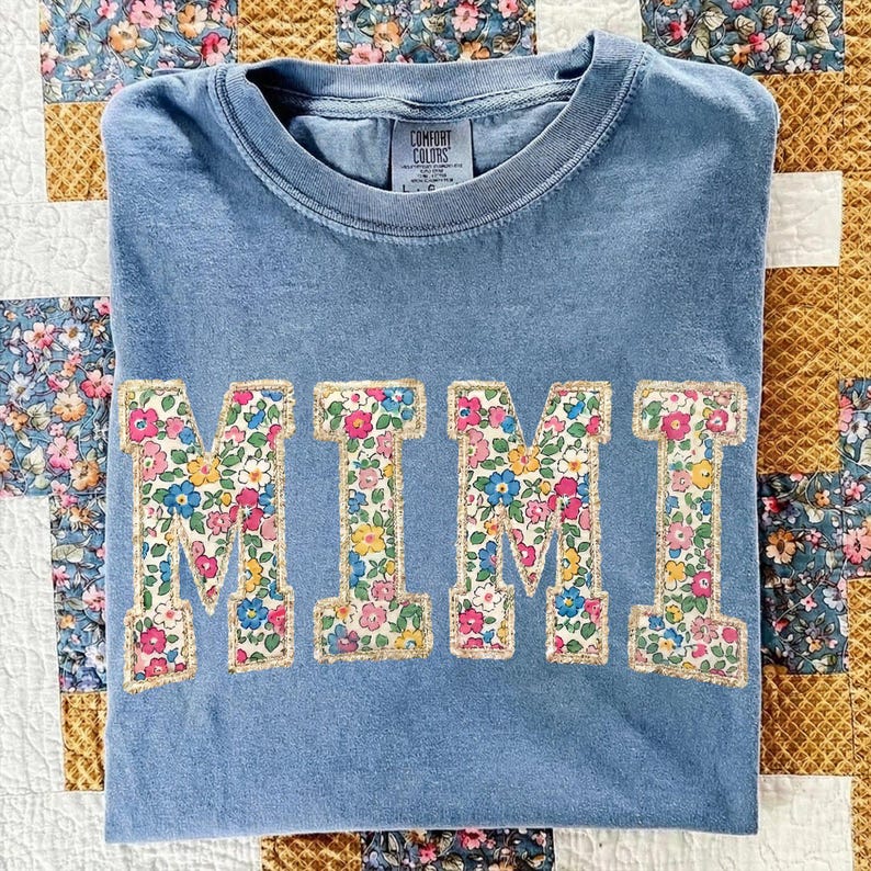 K&ouml;nnte beinhalten: Blaues Comfort Colors T-Shirt mit dem Wort "MIMI" in gro&szlig;en Buchstaben mit Blumenmuster und goldenem Rand. Das T-Shirt liegt gefaltet auf einem floralen Quilt. Ein l&auml;ssiges Kleidungsst&uuml;ck.
