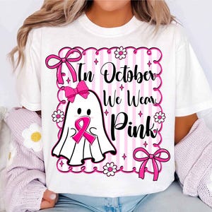 May include: White t-shirt with a pink and white design. The design features a ghost wearing a pink ribbon, surrounded by pink bows, flowers, and the text "In October We Wear Pink".