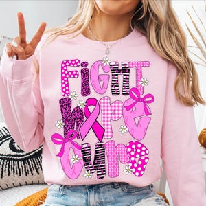 May include: Pink sweatshirt with the words "FIGHT MORE CURE" in a mix of patterns, including polka dots, leopard print, and stripes. The design incorporates pink ribbons and small white daisy accents, promoting breast cancer awareness.