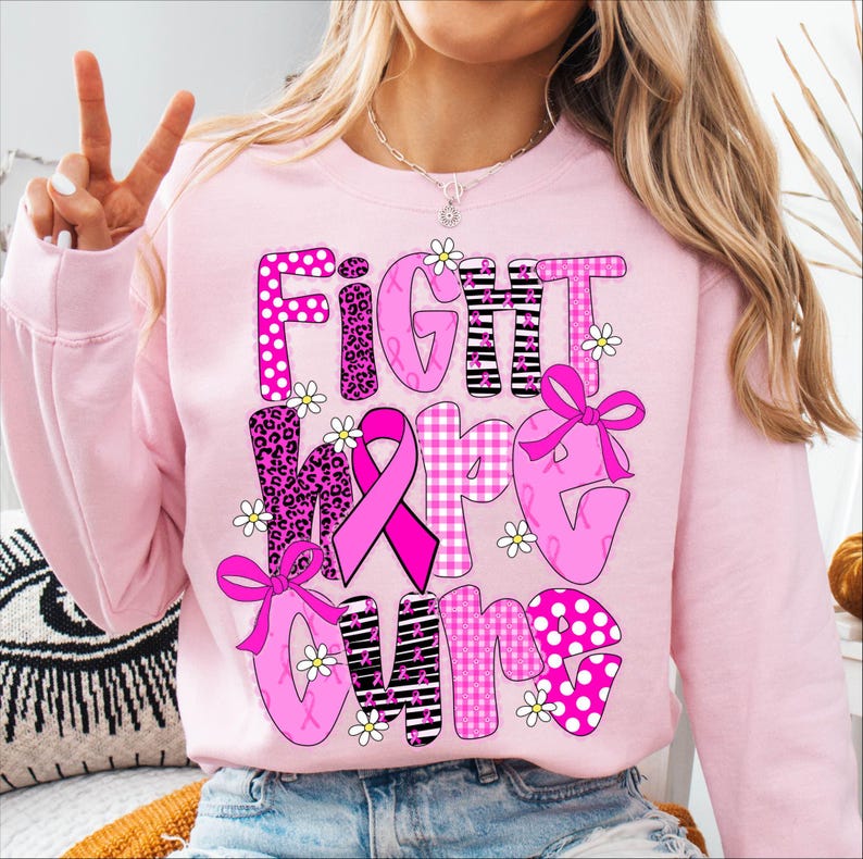 May include: Pink sweatshirt with the words "FIGHT MORE CURE" in a mix of patterns, including polka dots, leopard print, and stripes. The design incorporates pink ribbons and small white daisy accents, promoting breast cancer awareness.