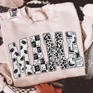 May include: A light beige crewneck sweatshirt featuring the word MAMA printed in large block letters. Each letter contains a different pattern: hockey pucks, a black and white checkerboard with hockey masks and sticks, leopard print, and ice skates.