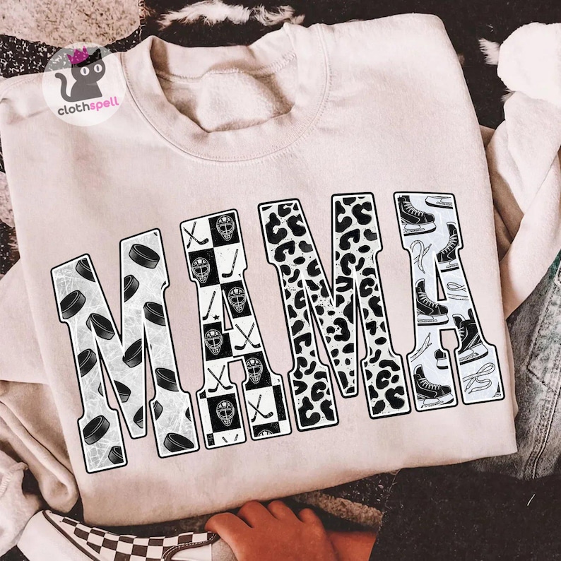 May include: A light beige crewneck sweatshirt featuring the word MAMA printed in large block letters. Each letter contains a different pattern: hockey pucks, a black and white checkerboard with hockey masks and sticks, leopard print, and ice skates.