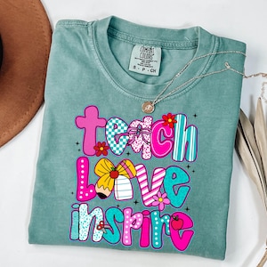 May include: A seafoam green t-shirt with the words "Teach Love Inspire" in a colorful, playful font. The letters are decorated with patterns, flowers, and a pencil. A gold necklace with a pendant is draped across the shirt.