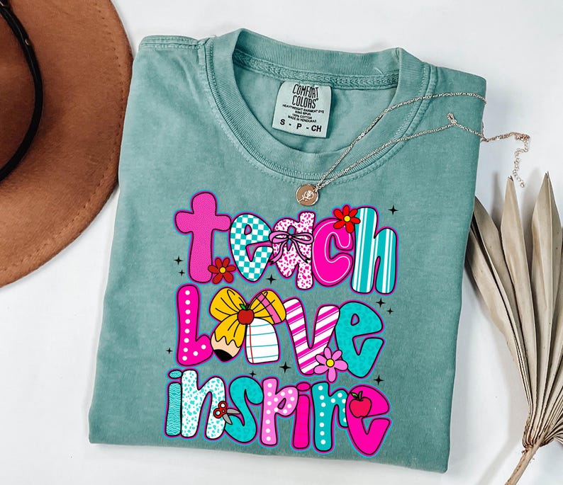 May include: A seafoam green t-shirt with the words "Teach Love Inspire" in a colorful, playful font. The letters are decorated with patterns, flowers, and a pencil. A gold necklace with a pendant is draped across the shirt.
