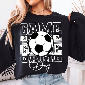 Może przedstawiać: Czarna bluza z białym graficznym piłką nożną i białymi słowami "GAME DAY". Słowo "GAME" jest ułożone w stos trzy razy. Piłka nożna jest czarno-biała.