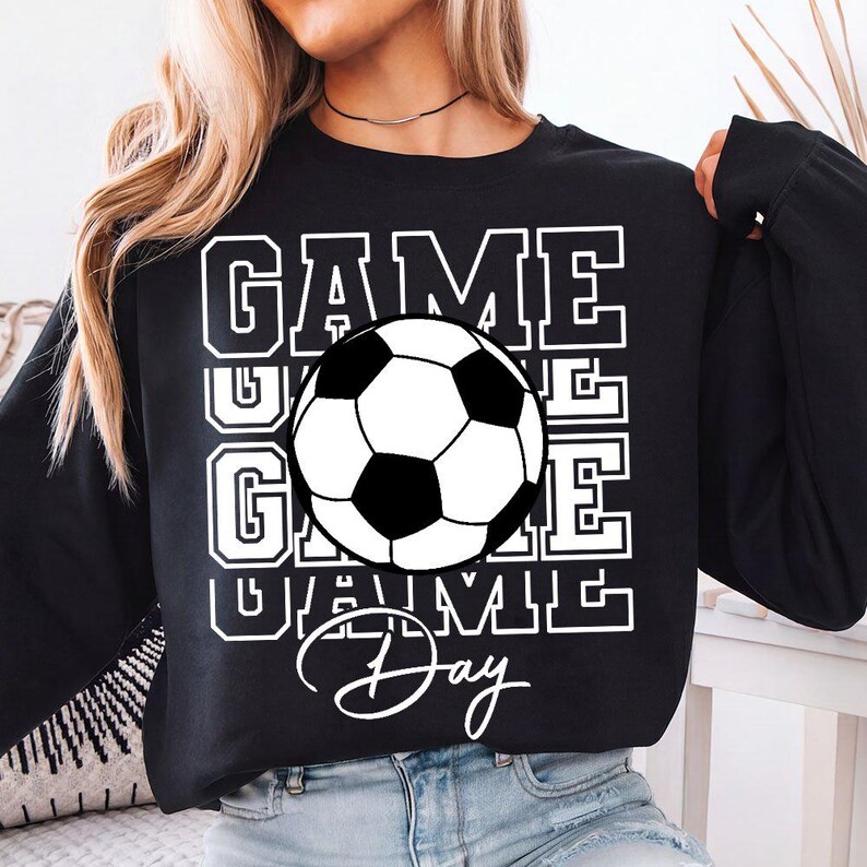 Może przedstawiać: Czarna bluza z białym graficznym piłką nożną i białymi słowami "GAME DAY". Słowo "GAME" jest ułożone w stos trzy razy. Piłka nożna jest czarno-biała.