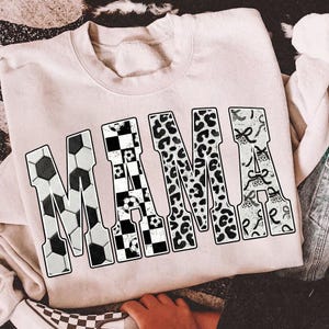 May include: A light pink sweatshirt with the word "MAMA" in large, bold letters. The letter "M" is designed with a soccer ball pattern, the "A" with a checkered pattern, the second "M" with a leopard print, and the final "A" with a decorative pattern.