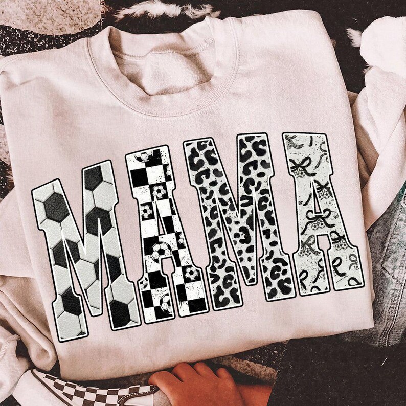 May include: A light pink sweatshirt with the word "MAMA" in large, bold letters. The letter "M" is designed with a soccer ball pattern, the "A" with a checkered pattern, the second "M" with a leopard print, and the final "A" with a decorative pattern.