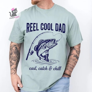May include: A pale green t-shirt featuring the text "REEL COOL DAD" above a navy blue illustration of a fish leaping from water with a fishing rod. The text "cast, catch & chill" is below the fish.
