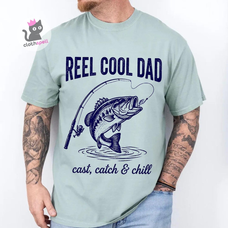 May include: A pale green t-shirt featuring the text "REEL COOL DAD" above a navy blue illustration of a fish leaping from water with a fishing rod. The text "cast, catch & chill" is below the fish.