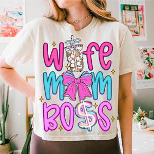 May include: Cream-colored t-shirt with the words "Wife Mom Boss" in pink and blue, accented with a coffee drink illustration and a pink bow. The design includes a dollar sign and star accents, creating a fun, casual look.