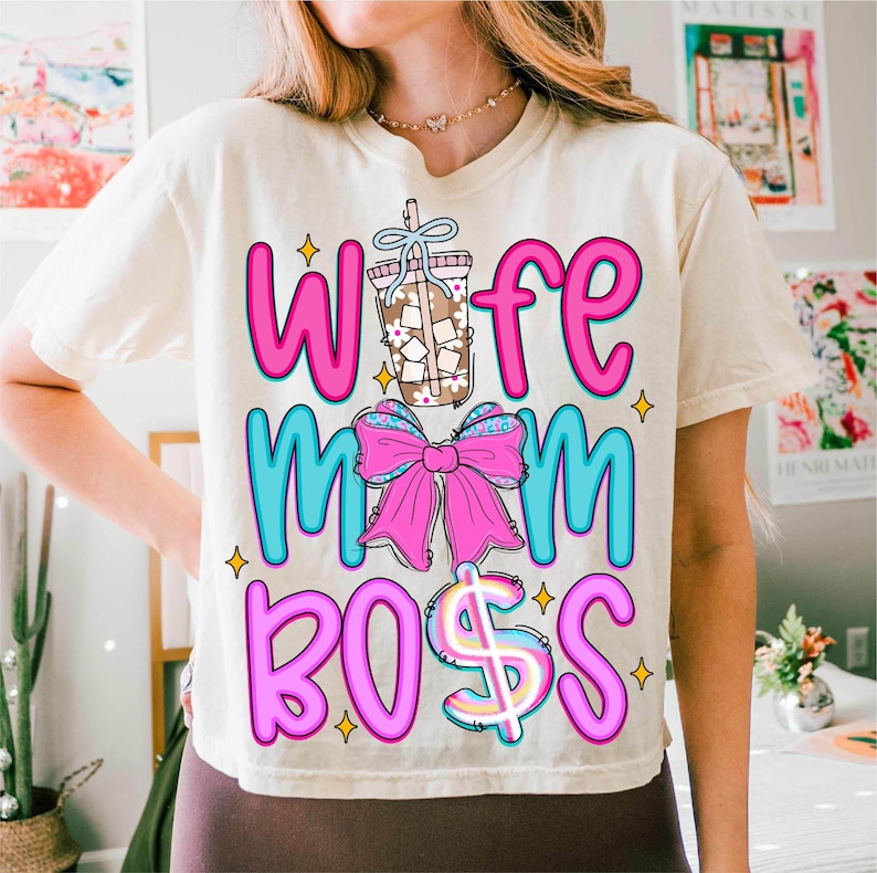 May include: Cream-colored t-shirt with the words "Wife Mom Boss" in pink and blue, accented with a coffee drink illustration and a pink bow. The design includes a dollar sign and star accents, creating a fun, casual look.