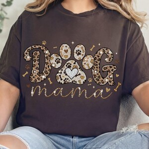 May include: A brown t-shirt with the words "DOG mama" in a decorative font. The word "DOG" is designed with a leopard print pattern and a paw print with a heart in the center. The word "mama" is in a cursive font.