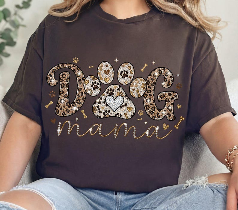 May include: A brown t-shirt with the words "DOG mama" in a decorative font. The word "DOG" is designed with a leopard print pattern and a paw print with a heart in the center. The word "mama" is in a cursive font.