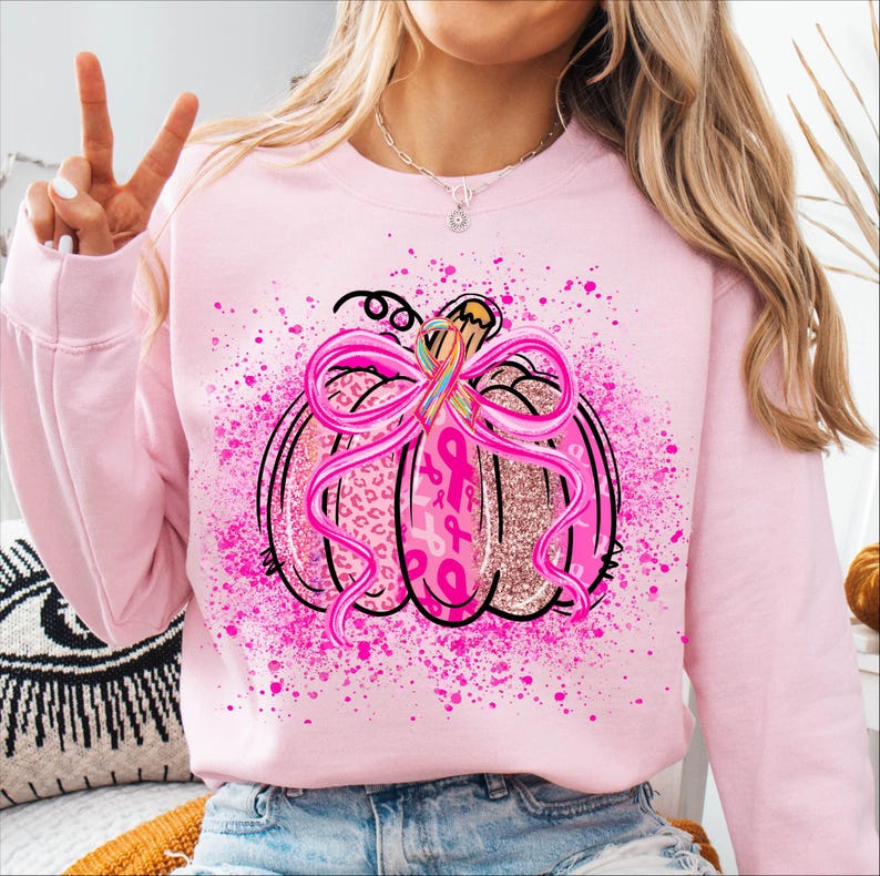 May include: Pink sweatshirt featuring a pumpkin design with a pink ribbon and breast cancer awareness ribbons. The pumpkin has a glittery texture and is outlined in black. Pink paint splatters surround the pumpkin.