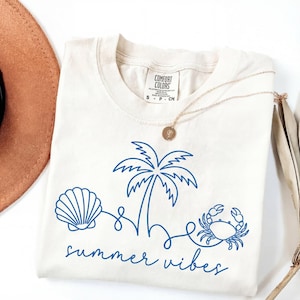 May include: Cream-colored t-shirt with a blue line drawing of a seashell, palm tree, and crab. The words "summer vibes" are written in a script font below the graphic. A gold necklace and a brown hat are partially visible.