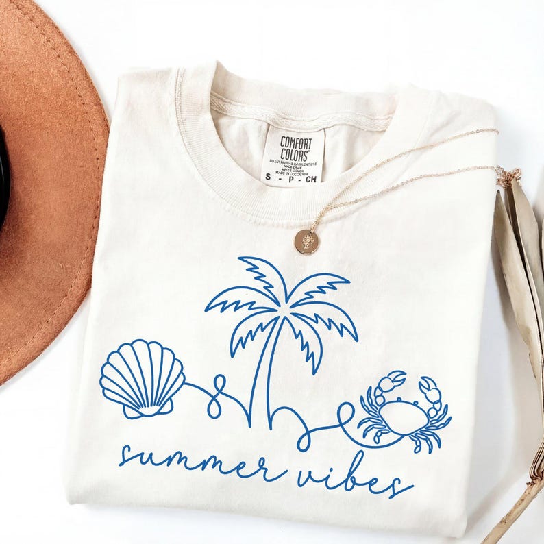 May include: Cream-colored t-shirt with a blue line drawing of a seashell, palm tree, and crab. The words "summer vibes" are written in a script font below the graphic. A gold necklace and a brown hat are partially visible.