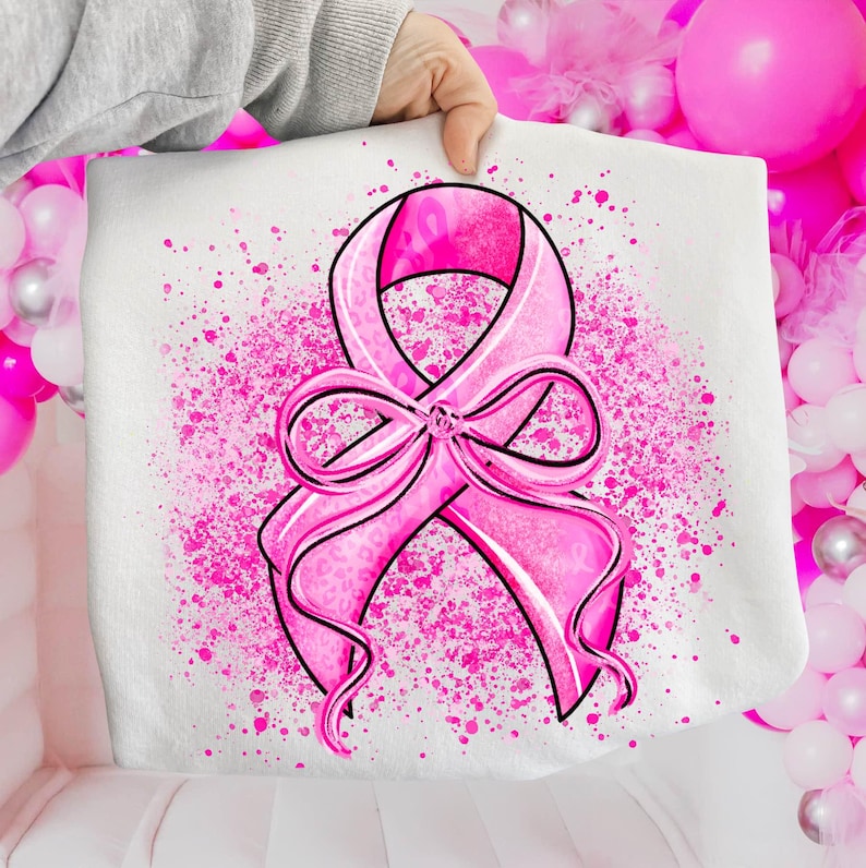 May include: A white tote bag featuring a large pink ribbon design, a symbol of breast cancer awareness. The ribbon is detailed with a leopard print pattern and outlined in black. Pink paint splatters surround the ribbon.