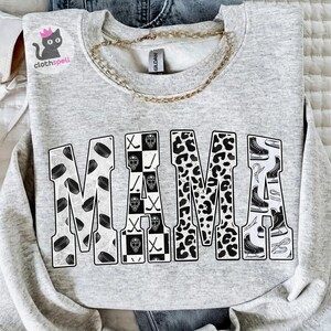 May include: A light grey crewneck sweatshirt featuring the word MAMA in large block letters. The letters are filled with black and white patterns including hockey pucks, a checkered hockey mask design, leopard print, and ice skates.